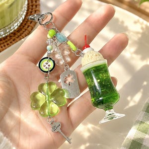 May include: A keychain featuring a miniature green drink charm with a cherry, a large translucent green four-leaf clover charm, a silver-toned key charm, a green button, a small silver tag with a clover cutout, and various small colorful beads.
