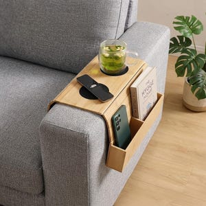 Puede incluir: Una bandeja de bambú para el brazo del sofá en color claro con un diseño flexible y articulado. Cuenta con un hueco circular para una taza de cristal, una superficie plana para un mando a distancia y un bolsillo lateral con un smartphone y un libro titulado The Hygge Home.