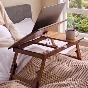 May include: A brown wooden laptop desk with an adjustable tilting surface and a flat side section. The desk features foldable legs and a metal support bracket for the tilt mechanism. A grey laptop sits on the tilted portion, and a beige mug rests on the flat side.