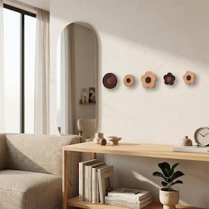 Puede incluir: Un conjunto de cinco ganchos de pared de madera en varias formas y tamaños, incluyendo diseños circulares y en forma de flor. Cada gancho presenta un exterior de madera clara con un centro circular marrón oscuro.