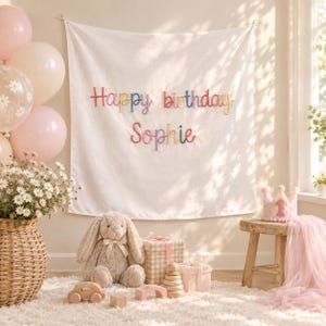May include: A white fabric wall hanging featuring the multicoloured embroidered text Happy birthday Sophie. The display includes light pink balloons, a bouquet of daisies in a woven basket, a plush rabbit, gift boxes, and a small pink crown.