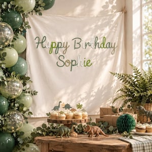 May include: A white fabric backdrop featuring the text Happy Birthday Sophie in green and white cursive lettering. The display includes a balloon arch in shades of green, dinosaur figurines, and cupcakes on a wooden stand, creating a festive scene.