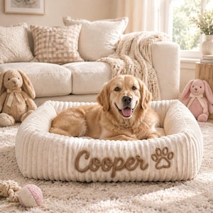 Pode incluir: Uma cama retangular para animais de estimação na cor creme com uma textura macia e canelada. A frente da cama apresenta o nome Cooper bordado em letras cursivas castanhas, acompanhado por um desenho de pata bordado em castanho à direita.