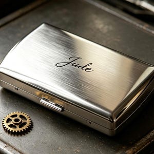 May include: A rectangular, brushed silver metal cigarette case featuring the name Jude engraved in a black script font on the top surface, accompanied by a small, circular, brass-colored gear component resting beside the case.