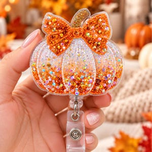May include: A pumpkin-shaped badge reel featuring a clear acrylic base covered in orange and white rhinestones. The design includes a prominent orange rhinestone bow at the top and a gold-toned stem. The reel has a clear plastic clip attachment.