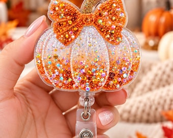 Op de afbeelding: Een pompoenvormige badgehouder met een doorzichtige acrylbasis bedekt met oranje en witte strass-steentjes. Het ontwerp bevat een opvallende oranje strass-strik aan de bovenkant en een goudkleurige steel. Voorzien van een doorzichtige clip.