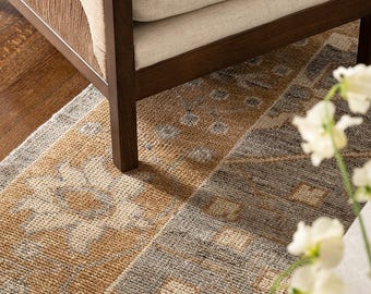 Pode incluir: Um tapete retangular com um padrão geométrico e floral repetitivo em tons de ocre, cinza-carvão e creme. O tapete está posicionado sob uma cadeira de madeira com assento estofado em creme e painéis laterais com textura.