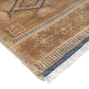 Pode incluir: Um tapete retangular com pelo texturizado em tons de castanho e bege. O tapete apresenta um padr&atilde;o geom&eacute;trico em azul suave e creme, com uma borda decorativa e franjas brancas ao longo da extremidade, exibindo uma constru&ccedil;&atilde;o tecida.