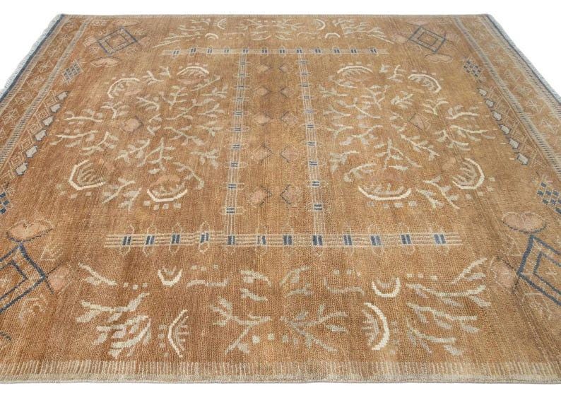 Pode incluir: Um tapete retangular grande em tom castanho claro quente com um padr&atilde;o sim&eacute;trico de formas geom&eacute;tricas, motivos florais estilizados e bordas lineares em branco e azul escuro. O design &eacute; tecido com pelo texturizado e extremidades com franjas.