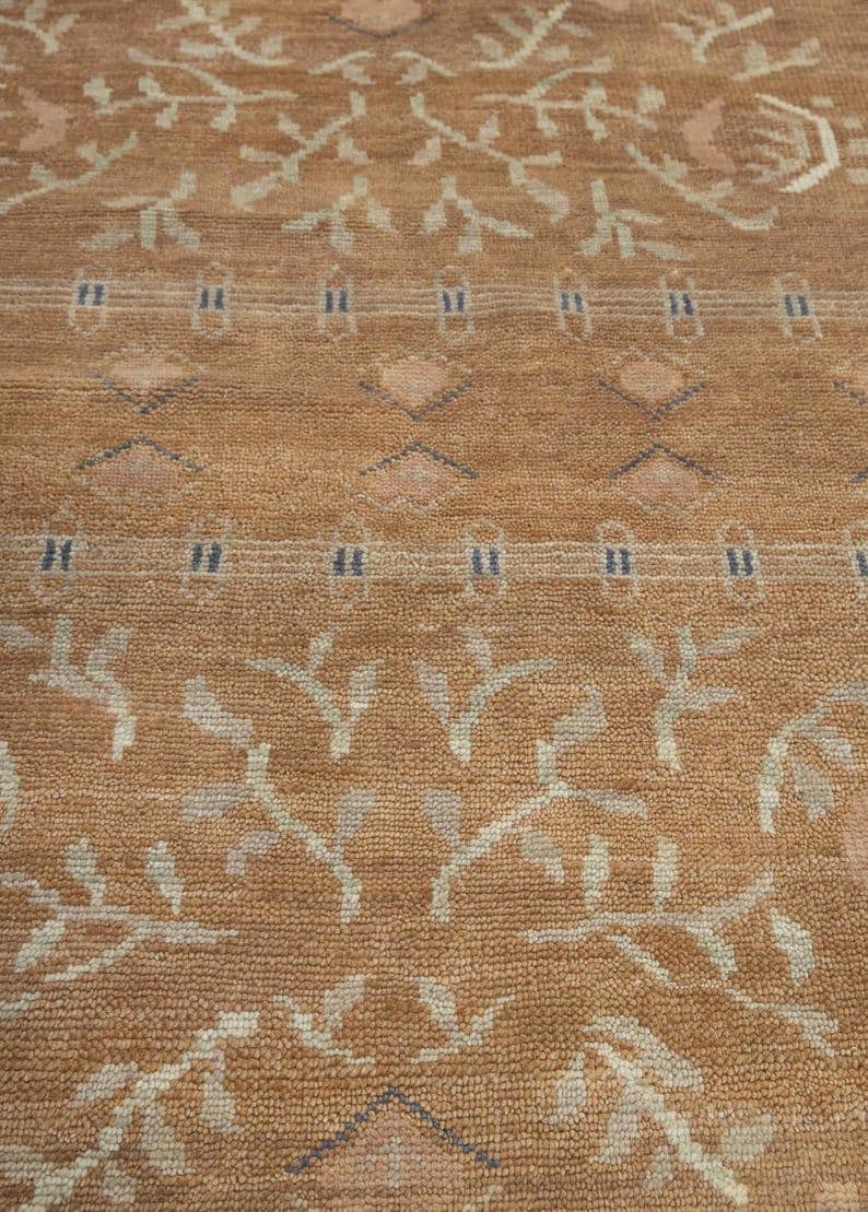 Pode incluir: Um tapete tecido com um fundo castanho claro quente e um padr&atilde;o repetitivo de ramos com folhas bege claro, pequenas formas de diamante e filas horizontais de tra&ccedil;os azuis finos. A textura parece densa e tufada com um pelo uniforme.