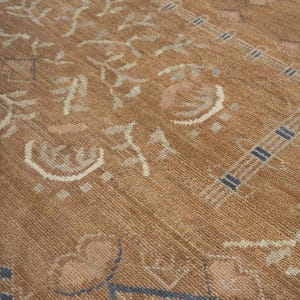Pode incluir: Um tapete castanho-claro com um padr&atilde;o repetitivo de formas geom&eacute;tricas, motivos florais estilizados e bordas lineares em creme e azul. O tapete tem uma superf&iacute;cie tecida e texturizada com uma altura de pelo uniforme.