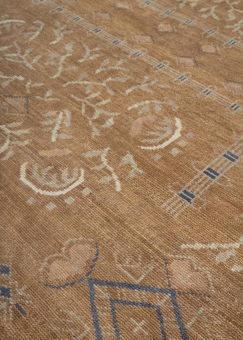 Pode incluir: Um tapete castanho-claro com um padr&atilde;o repetitivo de formas geom&eacute;tricas, motivos florais estilizados e bordas lineares em creme e azul. O tapete tem uma superf&iacute;cie tecida e texturizada com uma altura de pelo uniforme.