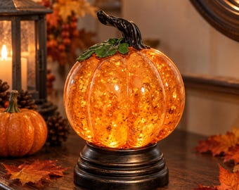 May include: A glowing orange glass pumpkin figurine with internal lights and gold-toned flecks, featuring a dark, curved stem and a green leaf accent. The pumpkin sits on a tiered, circular black base, creating a warm, illuminated display piece.
