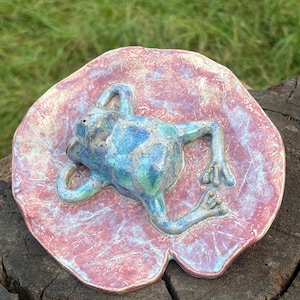 Könnte beinhalten: Eine Keramikskulptur eines Frosches, der auf einem Seerosenblatt ruht. Der Frosch ist in Blau- und Grüntönen glasiert, während das Seerosenblatt eine strukturierte, gesprenkelte Oberfläche in Rosa-, Rot- und Weißtönen mit sichtbarer Äderung aufweist.