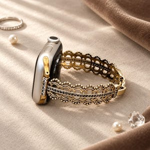 May include: A silver-toned smartwatch featuring a decorative gold-coloured metal band with an intricate, scalloped, lace-like pattern. The band is accented with a central row of small, dark, sparkling stones and connects to the watch casing with gold hardware.