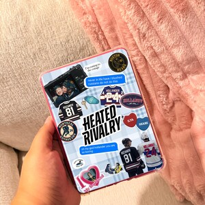 May include: A rectangular book cover featuring a light blue plaid background with numerous hockey-themed stickers. The text HEATED RIVALRY is centered in bold black letters. Various stickers include hockey jerseys, players, and speech bubbles with text.
