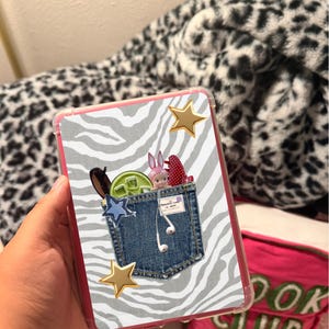 Op de afbeelding: Een rechthoekige tablethoes met een grijs en wit zebrapatroon op de achtergrond. Het midden toont een afbeelding van een spijkerbroekzak met daarin een klein roze konijnenfiguurtje, een groen rond voorwerp en witte bedrade oordopjes. Twee gouden sterren.