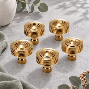 May include: A set of five circular cabinet knobs in a polished gold finish. Each knob features a series of concentric, raised horizontal ridges along the sides and a smooth, rounded centre dome, mounted on a short, cylindrical base.