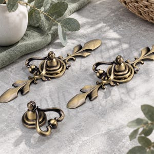 May include: Two elongated brass-toned metal drawer pulls with ornate, leaf-shaped ends and a central circular mounting base, accompanied by a single matching smaller circular knob featuring a curved, drop-style handle attachment.