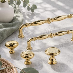 May include: A collection of polished gold-tone cabinet hardware including two long, curved drawer pulls with decorative center detailing and three distinct round knobs of varying shapes, all featuring a smooth, reflective metallic finish.