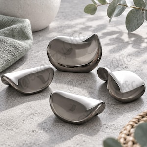 May include: Four small, smooth, metallic objects with a polished gunmetal finish. Each piece features a curved, bean-like shape with rounded edges and a reflective surface. The objects are arranged in a cluster, showcasing their uniform, sleek, and contoured design from various angles, highlighting the high-gloss, dark silver-toned metal construction.