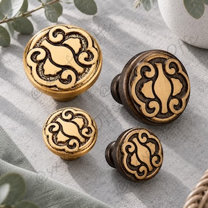 May include: Four round cabinet knobs in two sizes, featuring a raised central motif with swirling scrollwork patterns. Two knobs have a bright gold finish, while the other two feature a darker, antiqued bronze finish with recessed detailing.