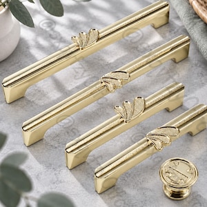 Puede incluir: Un juego de cuatro tiradores de armario de metal dorado y un pomo redondo a juego. Cada tirador presenta una barra rectangular larga con una franja central texturizada y una pieza decorativa en forma de abanico en la superficie superior.