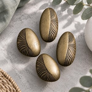 May include: Four oval-shaped cabinet knobs in a brushed brass finish. Each knob features a smooth, solid half-section and a textured half-section with radiating, curved lines that create a fan-like pattern across the surface of the metal hardware.
