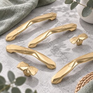 May include: A collection of five gold-toned cabinet hardware pieces, including three elongated handles and two smaller knobs. Each piece features a faceted, geometric surface with sharp angles and a polished metallic finish, creating a multi-faceted appearance.