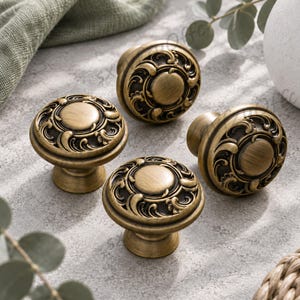 May include: A set of four round cabinet knobs in an antique brass finish. Each knob features a central circular disc surrounded by an intricate, raised scrollwork pattern. The knobs have a smooth, metallic texture with dark recessed areas.