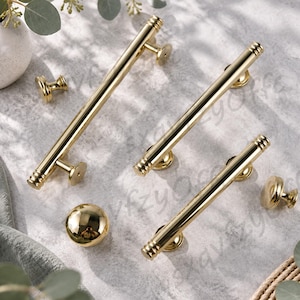 May include: A collection of gold-toned cabinet hardware including three cylindrical bar pulls with ribbed end details and two matching round knobs. Each piece features a polished metallic finish and circular mounting bases for attachment.