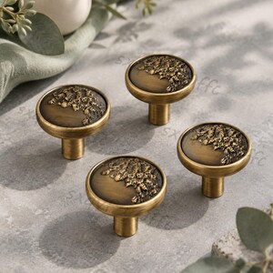 May include: Four round cabinet knobs with a brushed antique brass finish. Each knob features a flat circular face with a textured, raised relief pattern covering one side, while the remaining surface is smooth and dark, creating a contrast.