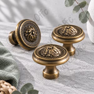 May include: Three round cabinet knobs with an antique brass finish. Each knob features a central raised floral relief pattern surrounded by a beaded border and a smooth outer rim. The knobs have a sturdy cylindrical base and a polished metallic surface.