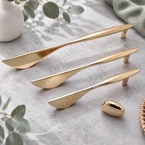 Könnte beinhalten: Ein Set aus drei goldfarbenen Metall-Schrankgriffen mit schlankem, länglichem und spitz zulaufendem Design sowie einem passenden ovalen Schrankknopf. Jedes Teil hat eine glatte, reflektierende Oberfläche und einen stabilen Befestigungsfuß.