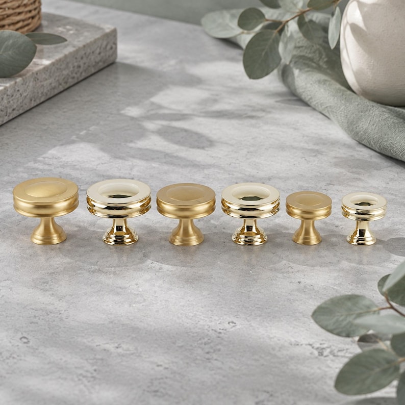 K&ouml;nnte beinhalten: Ein Set aus sechs goldfarbenen Schrankkn&ouml;pfen aus Metall, die in einer Reihe angeordnet sind. Die Kn&ouml;pfe haben eine kreisf&ouml;rmige Oberseite mit abgestuftem Rand und einen konischen Sockel. Einige Kn&ouml;pfe haben eine flache Oberseite.