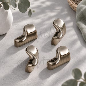 May include: A set of four metallic, gold-toned chopstick rests with a smooth, curved, and abstract sculptural form. Each piece features a reflective, polished surface with fluid, organic contours designed to support chopsticks horizontally.