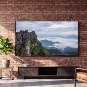 May include: A large flat-screen television mounted on a red brick wall displays a landscape image of mountains and clouds with the text DOI PHA TANG, CHIANG RAI, THAILAND. Below the screen is a dark brown wooden media console with open shelving.