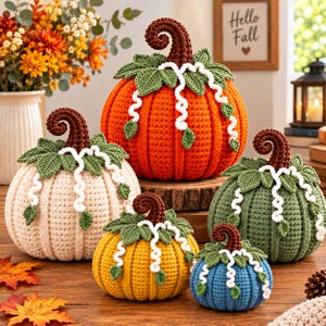 May include: A collection of five crocheted pumpkins in orange, cream, yellow, green, and blue. Each pumpkin features a dark brown curly stem, green crocheted leaves, and white decorative tendrils draped over the top of the ribbed surface.