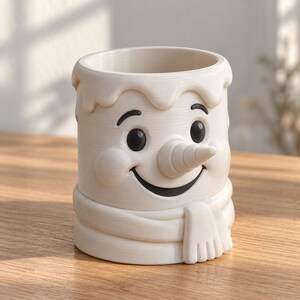 May include: A cylindrical off-white ceramic mug featuring a sculpted snowman face with a carrot-shaped nose, black eyes, a wide smiling mouth, and a textured scarf wrapped around the base. The top rim has a decorative dripping wax effect.