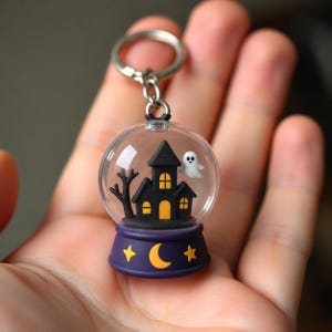 May include: A clear spherical keychain featuring a miniature black haunted house with yellow windows, a small white ghost, and a bare black tree inside. The base is dark purple with a yellow crescent moon and two yellow stars printed on the front.