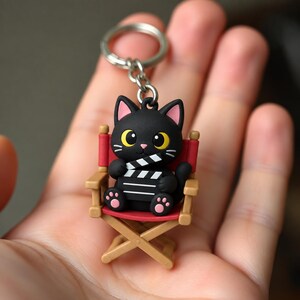 May include: A keychain featuring a black cat figurine with large yellow eyes and pink inner ears, sitting on a miniature red and tan director's chair. The cat is holding a small black and white film clapperboard in its paws, with pink paw pads.