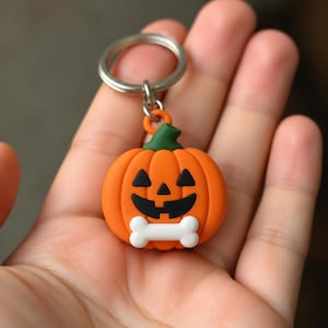 Może przedstawiać: Brelok z zawieszką w kształcie jasnopomarańczowej dyni z zieloną łodyżką, czarną twarzą jack-o'-lantern oraz białym elementem w kształcie kości u dołu. Zawieszka jest przymocowana do okrągłego srebrnego metalowego kółka na klucze i akcesoria.