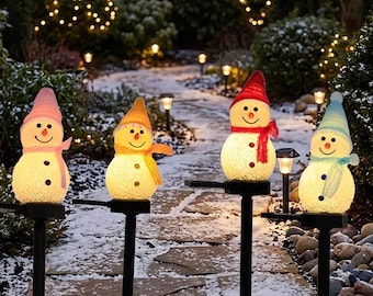 May include: Four illuminated snowman garden stakes with textured white bodies, black button details, and orange carrot noses. Each snowman wears a different colored hat and scarf: pink, yellow, red, and light blue, mounted on black poles.