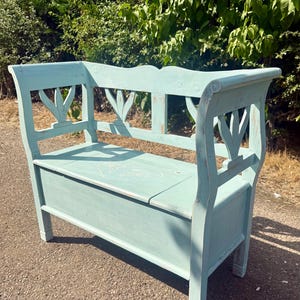 May include: A light blue wooden storage bench with a hinged seat lid. The backrest features three vertical cut-out details, and the seat surface displays a faint, etched heart design. The paint finish shows signs of wear along the edges.