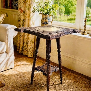 May include: A dark brown, intricately carved wooden side table with a square top and three legs. The table features a decorative shelf and a brass-coloured circular insert on the tabletop. The table is placed on a woven rug.