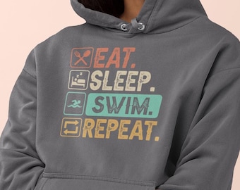 Op de afbeelding: Een donkergrijs hoodie met een vintage-stijl grafische afbeelding met de tekst "Eten. Slapen. Zwemmen. Herhalen."