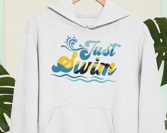 Op de afbeelding: Witte hoodie met een voorzak en de tekst "Just Swim" in een kleurrijk, gestileerd lettertype. Het ontwerp bevat golf- en waterafbeeldingen in tinten blauw, geel en zwart. De hoodie hangt aan een houten hanger.