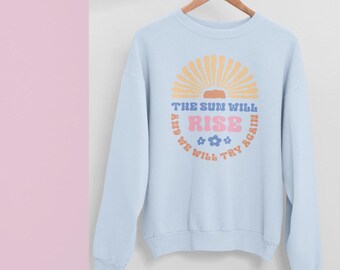 May include: Light blue sweatshirt with a retro design. The graphic features a sunburst, the word "RISE" in pink, and the text "THE SUN WILL RISE AGAIN AND WE WILL TRY AGAIN" in blue. The sweatshirt is hanging on a wooden hanger.