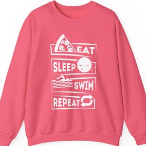 May include: Pink sweatshirt with a white graphic that says "Eat Sleep Swim Repeat" with icons of a pizza slice, a sleeping face, a swimmer, and a repeat symbol.