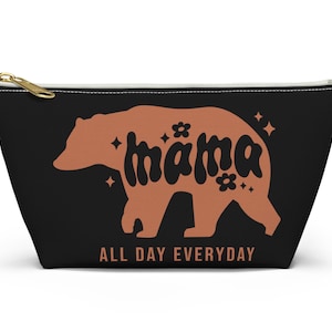 May include: Black zippered pouch featuring a brown bear silhouette. The bear is outlined with the word "mama" in a retro font. The text "All Day Everyday" is printed below the bear.